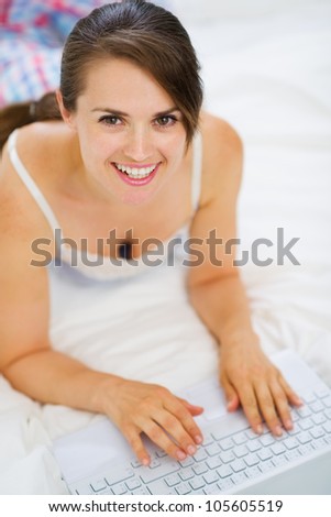 Smiling woman laying on bed and working on laptop. Upper view