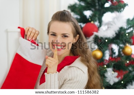 Smiling young woman put gift in Christmas socks and showing shh gesture