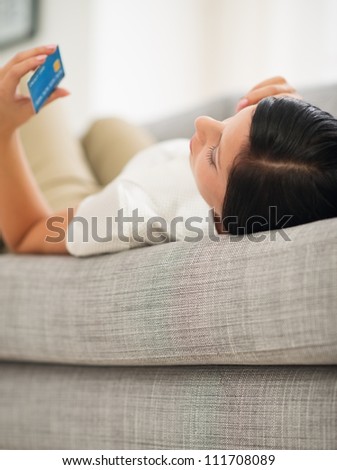 Woman laying on sofa speaking mobile and looking on credit card. Rear view