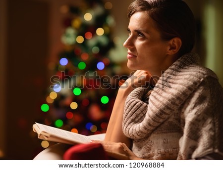 Dreaming young woman sitting chair and reading book in front of Christmas tree