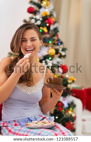 Happy young woman in pajamas eating cookies with hot chocolate near Christmas tree