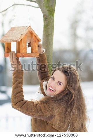 Smiling young woman hanging bird feeder on tree in winter outdoors