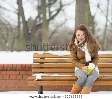 Happy young woman sitting on bench with hot beverage in winter outdoors
