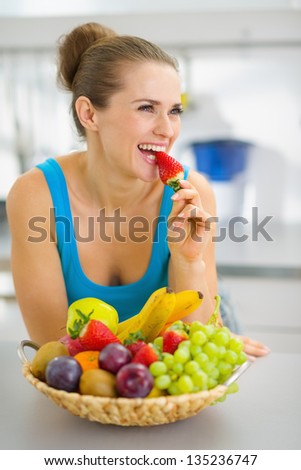 Happy young woman eating strawberry in modern kitchen