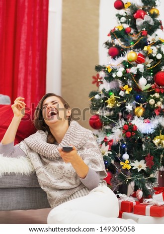 Laughing young woman with tv remote control near christmas tree