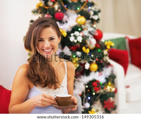 Happy young woman in pajamas near christmas tree drinking hot beverage