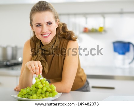 Happy young housewife eating grape in kitchen