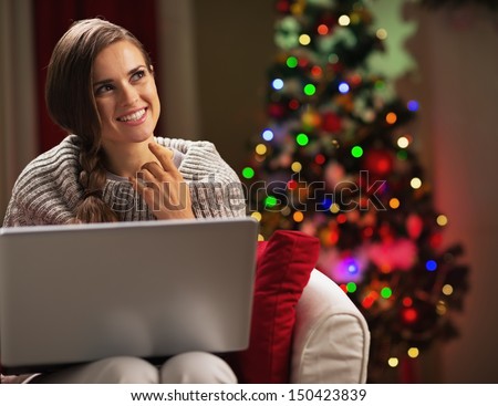 Smiling young woman with laptop near christmas tree looking on copy space