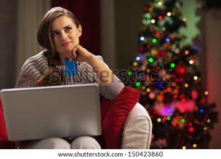 Thoughtful young woman with laptop and credit card near christmas tree