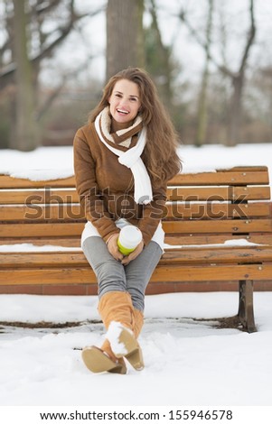 Happy young woman with cup of hot beverage sitting on bench in winter park