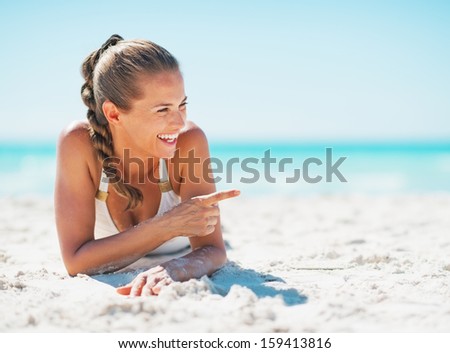 Smiling young woman in swimsuit laying on beach and pointing on copy space