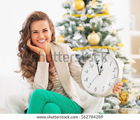 Happy young woman showing clock in front of christmas tree