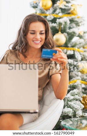 Portrait of happy young woman with credit card using laptop near christmas tree