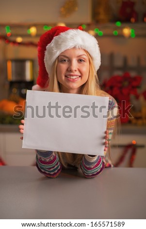 Happy teenage girl in santa hat showing blank paper sheet in christmas decorated kitchen