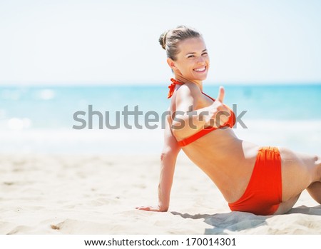 Smiling young woman sitting on beach and showing thumbs up