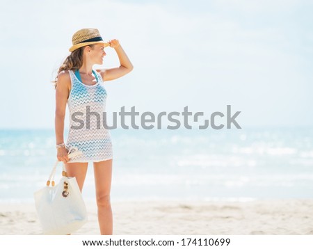 Full length portrait of young woman in hat and with bag on beach looking into distance