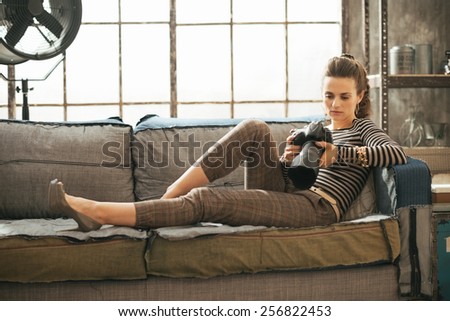 Happy young woman laying on sofa and using modern dslr photo camera