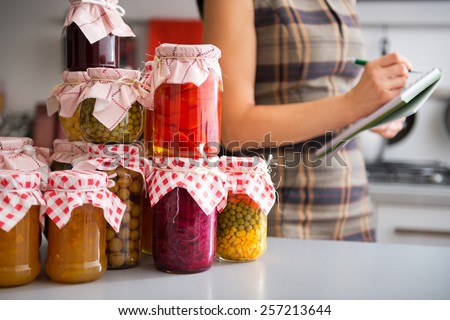 Closeup on jars with homemade fruits jam and pickled vegetables and housewife writing in notepad in background