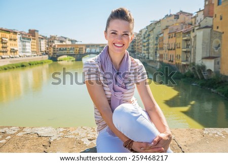 Portrait of happy young woman sitting on bridge overlooking ponte vecchio in florence, italy