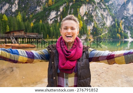 A brunette wearing outdoor gear is laughing and smiling while taking selfie. She is on the shores of Lake Bries. Behind her, wooden boats, a rustic pier, and golden and green trees reflect the autumn.