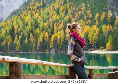 A brunette hiker wearing outdoor gear stands thinking on the shores of Lake Bries. The still water, golden colours and shades of gray provide an autumn background that is reflected in the still water.