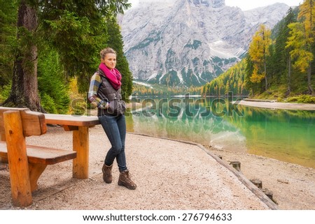 A brunette woman hiker wearing outdoor gear stands resting against a wooden picnic table along the edge of Lake Bries.