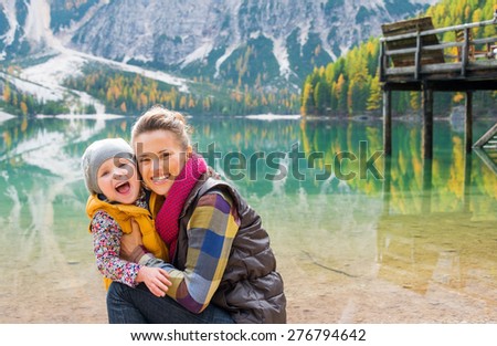 A kneeling mother and standing child wearing outdoor gear are cuddling, smiling and laughing on the shores of Lake Bries.