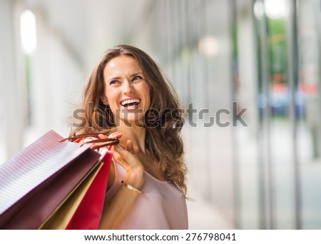 A brown-haired woman holding three shopping bags - brown, gold, and red - over her right shoulder looks back at someone who is making her laugh. A good shopping spree makes any woman happy.