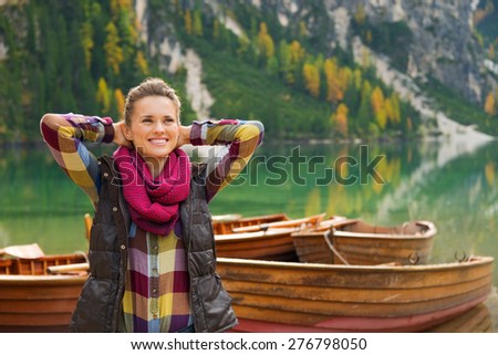 Wearing outdoor gear, a brunette woman hiker relaxes, hands behind her head. She is happy and content. In the background, wooden boats float on the water.