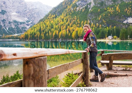A smiling brunette hiker wearing outdoor gear stands facing the water and leaning against a wooden railing. The still water in the background provides a perfect mirror image of the mountains and trees