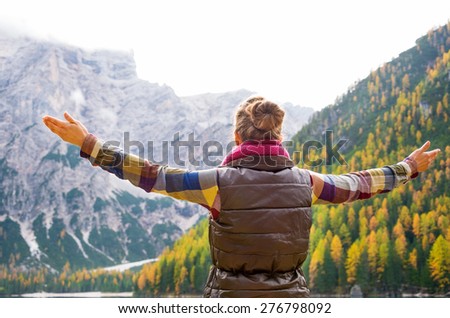 Seen from behind, a woman brunette hiker holds her arms wide open in joy. In the background, autumn leaves and the Dolomite mountains.