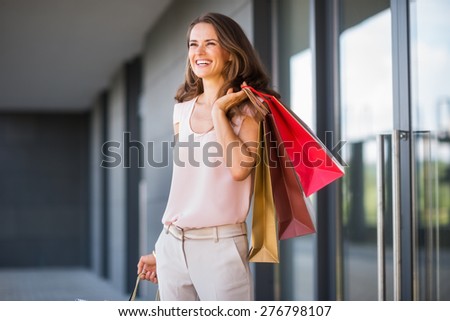 A brown-haired woman holding three shopping bags - gold, brown, and red - over her left shoulder laughs and smiles as she looks out into the distance. She is relaxed, and effortlessly stylish.
