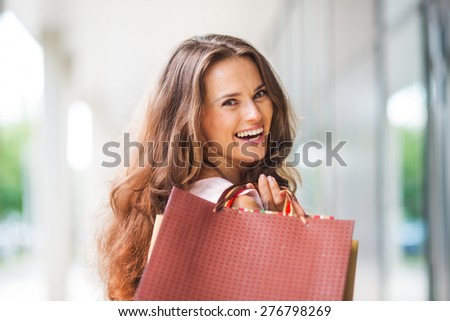 A brown-haired woman holding shopping bags - with a brown, textured bag most prominent - over her right shoulder looks back at the viewer in total happiness.