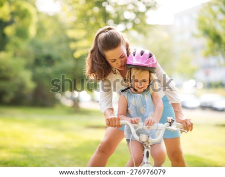 An encouraging mother helps her daughter learn how to steer her new bicycle. Wearing a pink helmet, the blonde daughter is proud and happy, looking down while smiling.