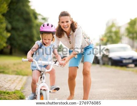 A laughing, smiling mother pushes her daughter forward on a warm summer\'s day as she teaches her how to ride her bicycle on a city sidewalk near a green park.