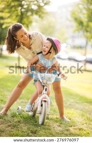 A mother hugs her daughter from behind, as daughter wearing a pink helmet looks up towards her mother, laughing and proud. She has just learned to ride her bike by herself, and is proud of herself.