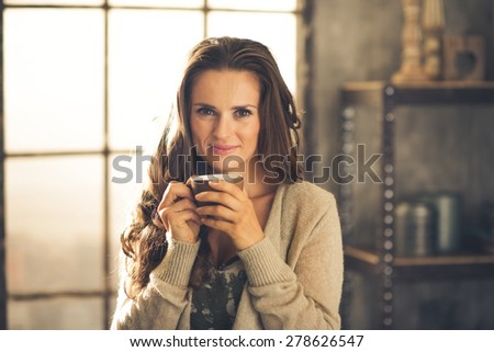 Head and shoulder shot of a brunette woman in comfortable clothing is smiling gently over the top of her coffee cup. She is casually dressed and in a cozy loft atmosphere. Urban chic decoration.