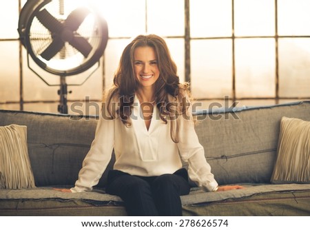 An elegant brunette is sitting a sofa in a loft, leaning forward and smiling. In the background, light is shining through a large window, industrial fan. Industry chic.
