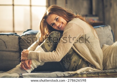 An elegant brunette woman is smiling, hugging her knees. Wearing comfortable, casual clothing, leggings, and a cardigan, she is relaxing on a loft sofa. Industrial chic ambiance and cozy atmosphere.