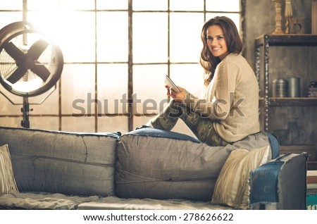 A brunette woman sitting on a sofa back, she is looking over her shoulder and smiling. Wearing comfortable clothing, she is holding a tablet pc. Industrial chic ambiance and cozy atmosphere.