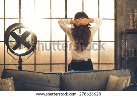 Elegant brunette woman seen from behind, leaning against the back of a sofa in a loft. Hands laced behind her head, she is looking out the loft window. Industrial chic atmosphere.