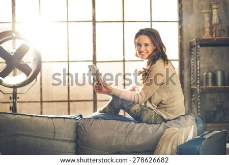 A brunette woman is smiling, looking up from her  tablet PC while sitting on the back of a sofa. Industrial chic ambiance and cozy atmosphere, sunlight is streaming through the loft window.