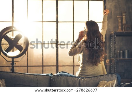 Seen from behind,a brunette woman is cheering, one arm raised, while talking on the phone. Industrial chic ambiance and cozy atmosphere, sunlight is streaming through the loft window.