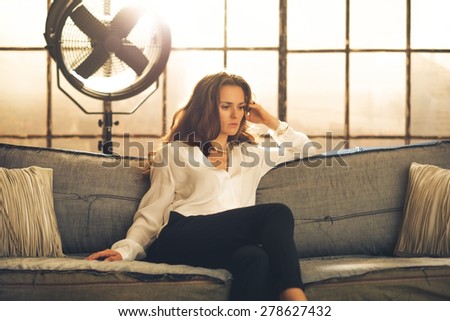 An elegant brunette is sitting on a sofa in a loft, pensive. In the background, a large window letting in light and an industrial standing fan.