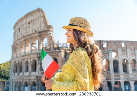 A woman tourist in summer is smiling and looking into the distance while holding an Italian flag. Behind her, the Colosseum.