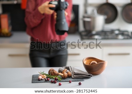 A woman photographer is taking a close-up of autumn fruits and vegetables - mushrooms, garlic, rosemary, and cranberries. On the counter, a wooden bowl. A gas stove and pot in the background.