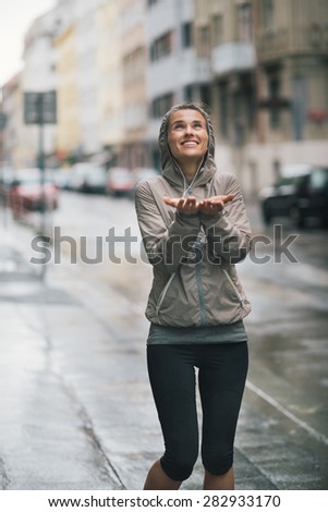 An athletic woman in workout and rain gear is looking up at the sky smiling, holding her hands out playfully to catch some rain drops...