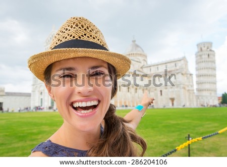 A smiling female tourist is pointing behind her to the Piazza dei Miracoli and the Leaning Tower of Pisa.