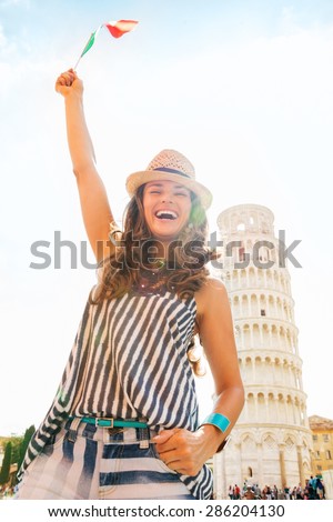 A beautiful, happy woman tourist laughs as she is waving the Italian flag up in the air. Behind her, the Leaning Tower of Pisa and crowds of tourists.
