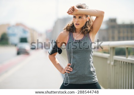 A woman jogger is taking a break, wiping sweat from her forehead with her forearm. She\'s taking a moment to catch her breath before getting down to the beat of her music and continuing her power run.